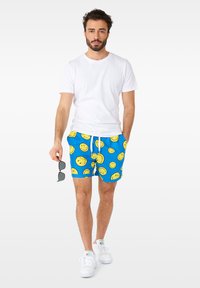 Shorts de bain bleus avec imprimé visage souriant jaune, dotés d'une taille élastique avec cordon de serrage. Portés avec un t-shirt blanc et des lunettes de soleil.