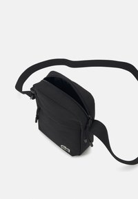 Bolso de hombro de tela negra con cierre de cremallera. Presenta un bolsillo frontal, correa ajustable y un pequeño logo bordado. Forma compacta y rectangular.