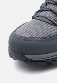 Κοντινή λήψη της μύτης ενός γκρι αθλητικού παπουτσιού με επάνω μέρος από δίχτυ, ενισχυμένο ράψιμο και υφασμένο καουτσούκ στην σόλα πάνω σε λευκό φ όντο.