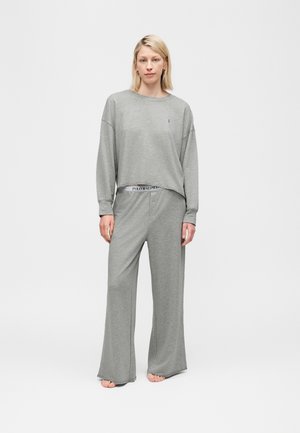 Γυναίκα όρθια, ξυπόλυτη, φορώντας φαρδιά γκρι μπλούζα με μακριά μανίκια και παντελόνι φαρδύ, με ορατή τη ζώνη Polo Ralph Lauren, σε απλό φόντο.