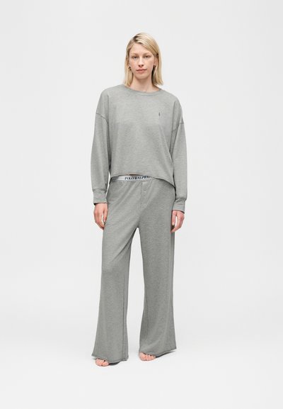 Femme debout pieds nus portant un haut ample à manches longues gris et un pantalon large avec une ceinture Polo Ralph Lauren visible, sur un fond uni.