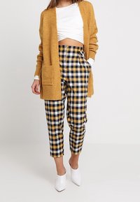 Sinapinkeltainen neulottu neuletakki, jossa avoin etuosa, yhdistetty mustiin ja keltaisiin ruutukuosiin leikattuihin housuihin. Asun kruunaavat valkoiset korolliset kengät.