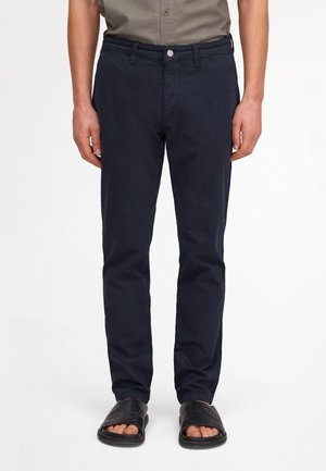 Mand iført marineblå slim-fit bukser, olivengrøn kortærmet skjorte stukket ind og sorte åbne sandaler, stående mod en hvid baggrund.