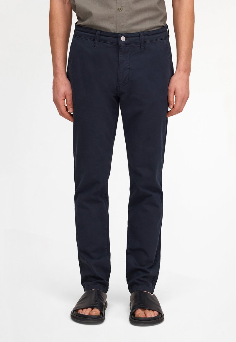 Homme portant un pantalon slim bleu marine, une chemise à manches courtes olive rentrée et des sandales noires ouvertes à l’avant, debout devant un fond blanc.