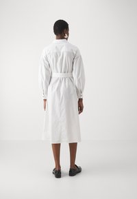 Abito camicia bianco a maniche lunghe con vita aderente, polsini abbottonati e orlo di lunghezza media. Tessuto liscio con dettaglio della vita con cintura.