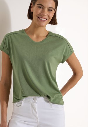 Femme souriante aux cheveux bruns courts porte un haut vert à manches courtes et un pantalon blanc, debout avec une main sur la hanche.