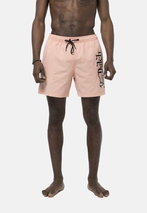 Modèle masculin portant un short de bain rose clair avec le logo noir "Von Dutch" sur la jambe droite, debout pieds nus devant un fond uni.