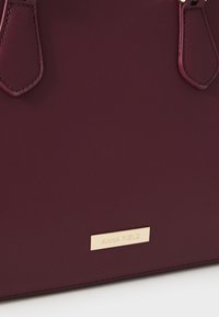 Borsa bordeaux con superficie testurizzata, dotata di due manici scuri e di una placca del marchio dorata. Design elegante e strutturato con hardware minimale.