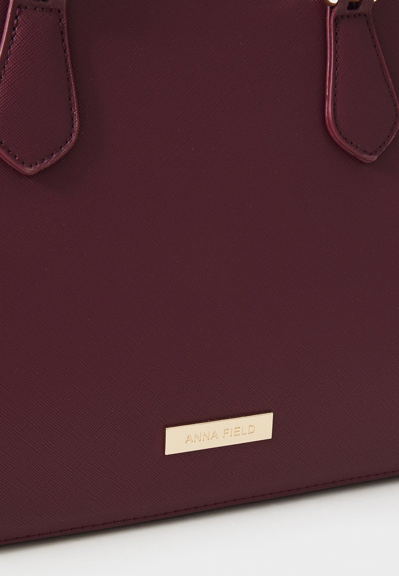 Bolso burdeos con superficie texturizada, que cuenta con dos asas oscuras y una placa de marca dorada. Diseño elegante y estructurado con hardware mínimo.