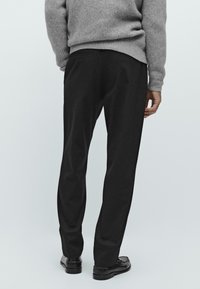 Man in zwarte broek, zwarte leren schoenen en een grijze gebreide trui, staand tegen een plain witte achtergrond, achterkant.