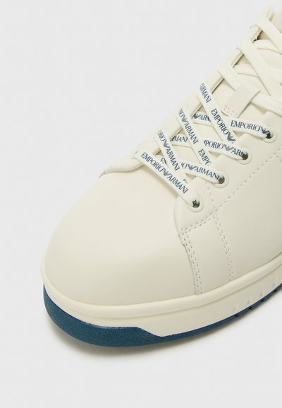 Sneaker en cuir blanc avec des accents bleus, présentant un bout rond, des lacets en textile avec logo et une semelle en caoutchouc texturée.