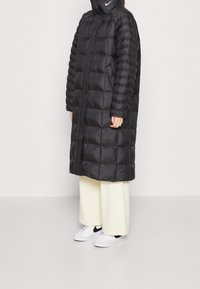 Lång svart puffjacka med hög krage, som har kviltade sektioner. Kombinerad med krämfärgade vida byxor och vita sneakers.