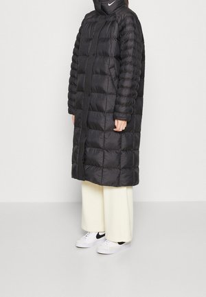 Abrigo puffer largo negro con un cuello alto, con segmentos acolchados. Combinado con pantalones de pierna ancha color crema y zapatillas blancas.