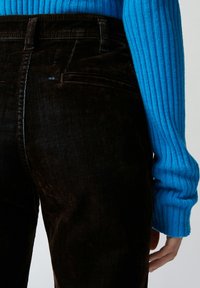 Crne kaderske hlače s glatkom teksturom, koje imaju stražnje džepove i uski kroj. Plavi rebrasti pulover djelomično je vidljiv.