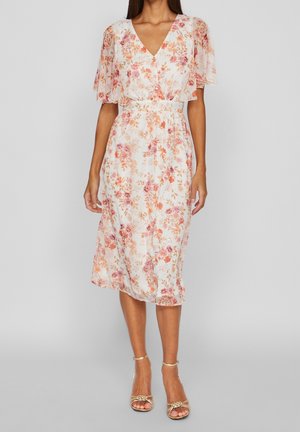 Kvinde iført en hvid midi-kjole med orange og pink blomsterprint og flæseærmer, kombineret med guld farvede sandaler med høje hæle og stropper.