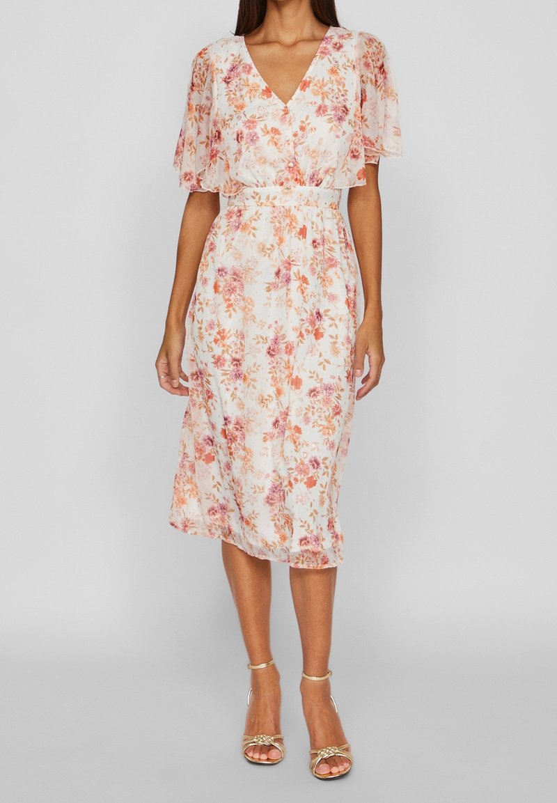Femme portant une robe midi blanche à imprimé floral orange et rose avec des manches volantées, associée à des sandales à talons hauts dorées à brides.