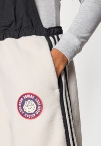 Φλις παντελόνια με κρεμ βάση, μαύρη ζώνη μέσ�ης και πλευρικές ρίγες. Διαθέτουν έναν στρογγυλό κόκκινο λεκέ με καρικατουρίστικο χαρακτήρα και κείμενο.