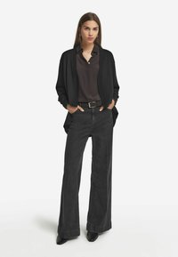 Svart cardigan över en mörk skjorta med knappar, parat med gråjeans med hög midja och vida ben. Svarta ankelskor fulländar looken.