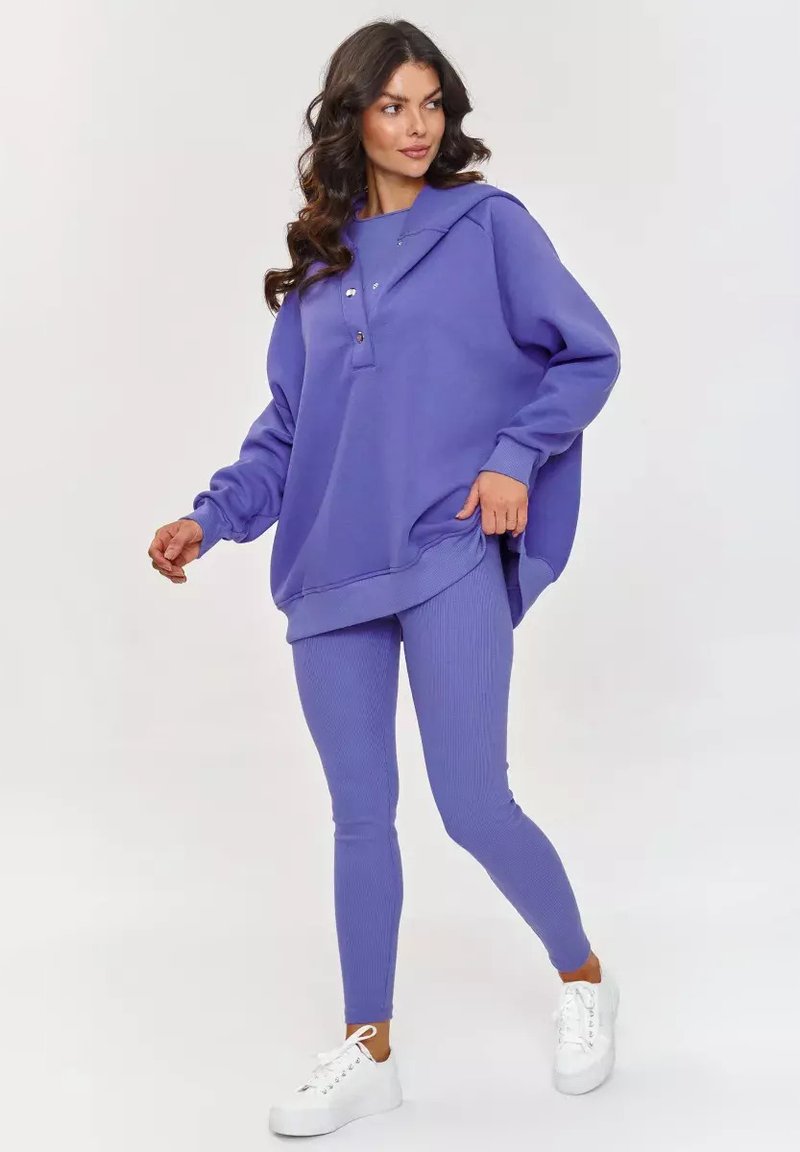 Fioletowa, oversize bluza z kapturem, z guzikami oraz fioletowe legginsy z prążkowanej tkaniny. Połączone z białymi trampkami, tworząc spójną stylizację.