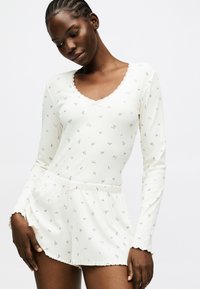 Γυναίκα που φοράει λευκό μακρυμάνικο τοπ και σορτς με μικρό μπλε λουλουδάτο σχέδιο, ποζάροντας με το κεφάλι σκυμμένο και τα χέρια χαλαρά.