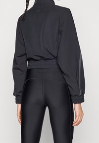 Osoba v černé krátké bundě a černých legínách s vysokým pasem, zobrazena zezadu, vlevo je vidět cop.