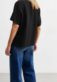 Černý texturovaný top s krátkými rukávy spojený s volnými modrými džínami s kapsami a pohodlným střihem. Prostředí je neutrální a minimalistické.