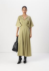 Rochie verde măsliniu, până la genunchi, cu decolteu adânc în V și mâneci largi. Țesătură texturată, talie strânsă. Asortată cu o poșetă neagră și cizme cu toc.