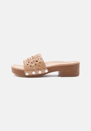Sandale beige à bout ouvert avec bride tressée, talon bloc en bois et clous argentés le long du bord de la semelle sur fond blanc.