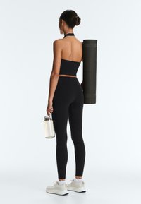 Žena v černém sportovním oblečení drží lahev na vodu a přes rameno nese srolovanou jóga podložku, otočená směrem od nás na bílém pozadí.