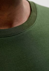 Camiseta de algodón verde con cuello redondo. La tela tiene una textura suave y el escote presenta detalles de costura visibles.