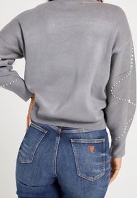 Grauer Strickpullover mit langen Ärmeln, besetzt mit Perlen entlang der Manschetten. Kombination mit blauen Jeans aus Denim mit aufgesetzten Taschen.