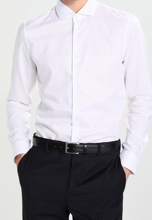 Mężczyzna ubrany w białą, długorękawową koszulę zapinaną na guziki, włożoną do czarnych spodni z czarnym skórzanym paskiem, z rękami w kieszeniach.