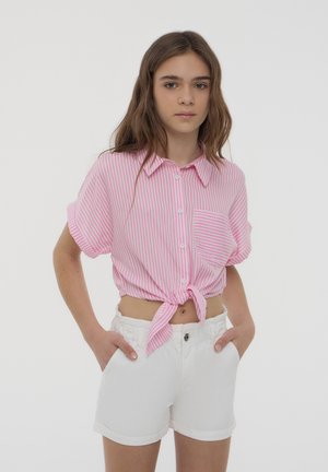Camisetta a maniche corte, a righe rosa chiaro e bianche, con un nodo sul davanti, dotata di colletto e taschino sul petto. Abbinata a pantaloni corti bianchi.