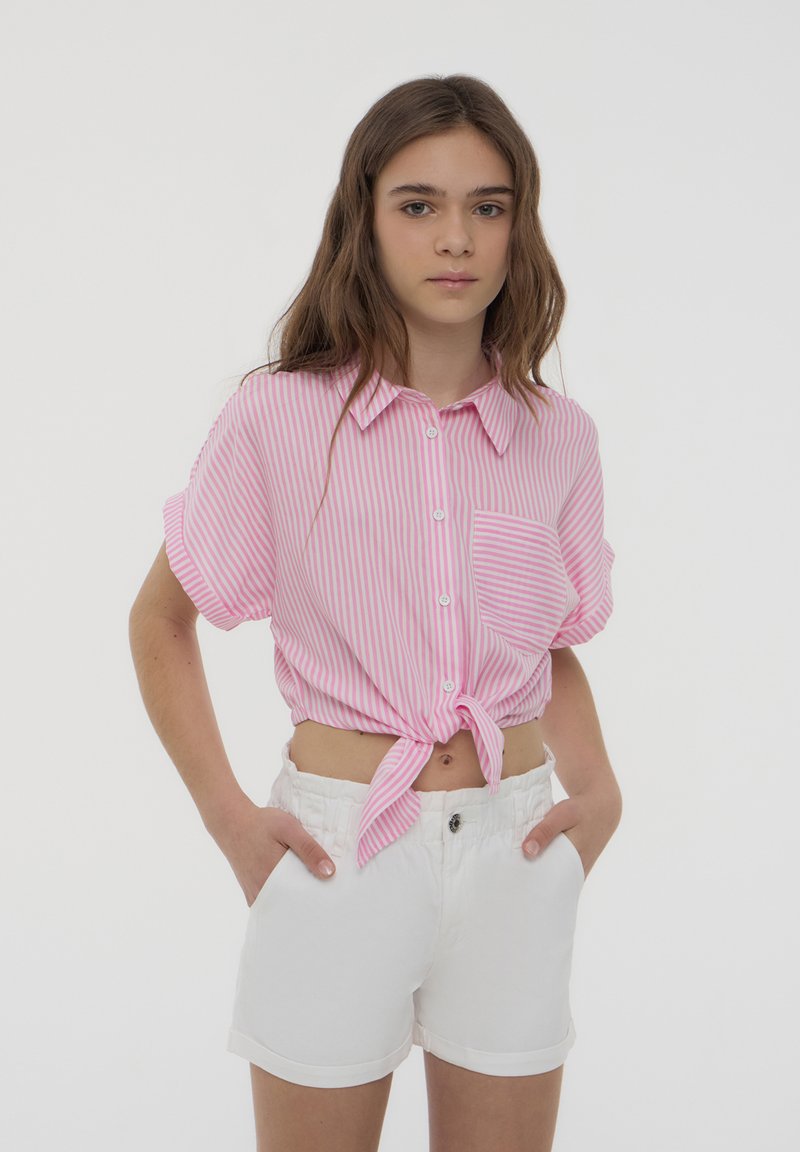Camisetta a maniche corte, a righe rosa chiaro e bianche, con un nodo sul davanti, dotata di colletto e taschino sul petto. Abbinata a pantaloni corti bianchi.