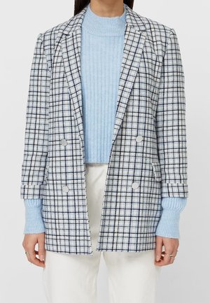 Parte superior del cuerpo de una persona que lleva un jersey azul claro de canalé debajo de una americana gris de cuadros cruzada y pantalones blancos.