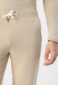 Beige joggebukser med ribbestrikket midjebånd, som har snor med metallspisser og sidelommer. Mykt, elastisk stoff med en glatt tekstur.