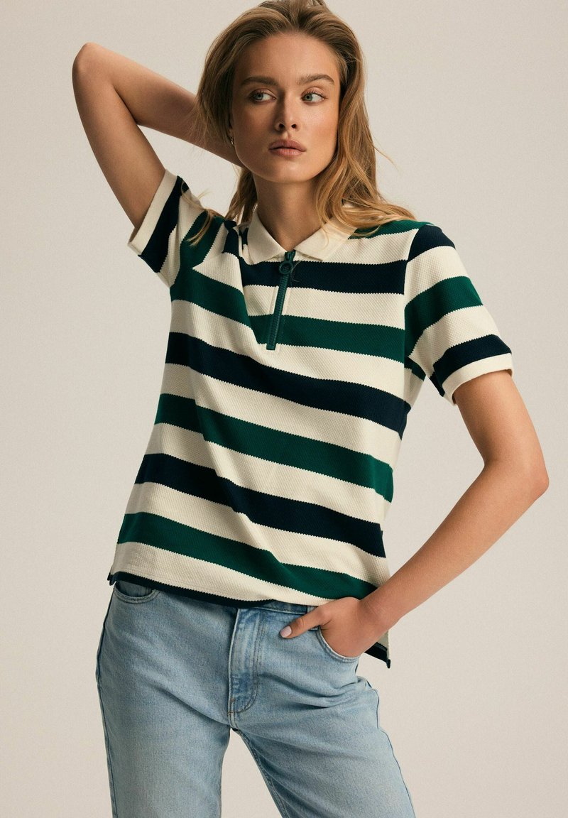 Koszulka polo w zielone, granatowe i kremowe poziome paski; z prążkowaną fakturą i kołnierzykiem; posiada częściowy zamek błyskawiczny; zestawiona z jasnoniebieskimi dżinsami.
