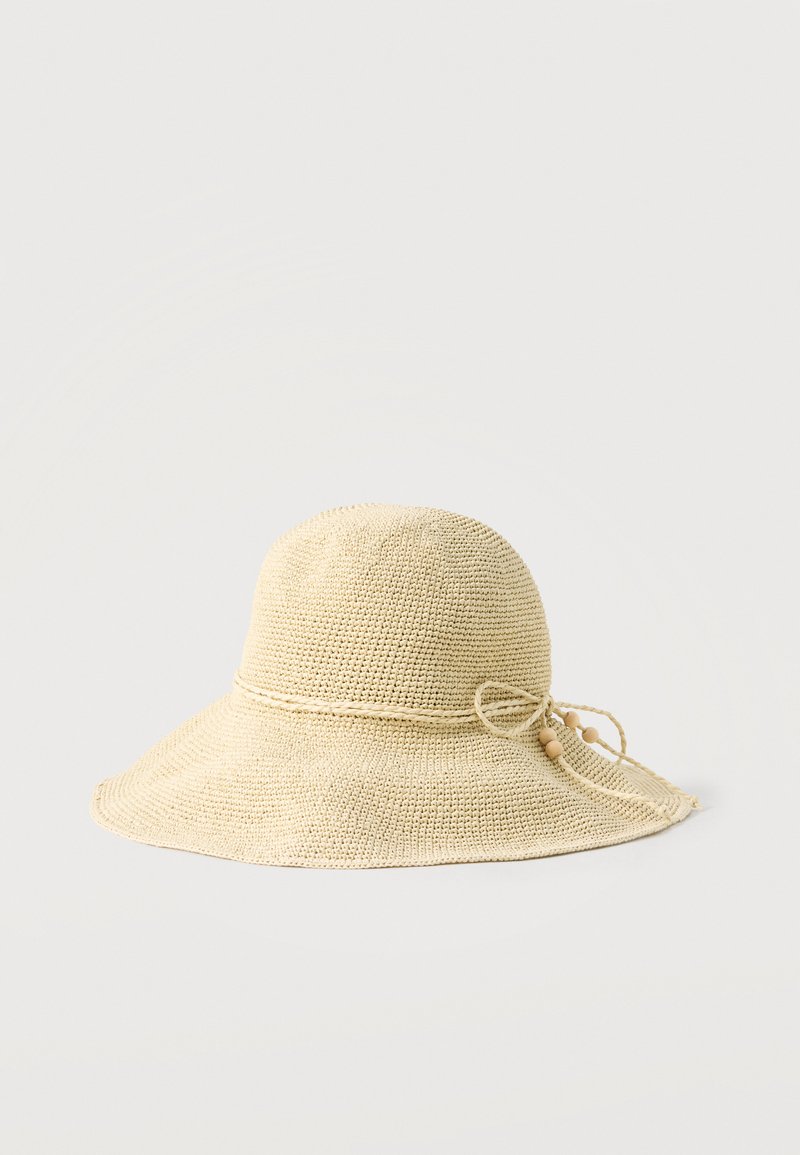 Chapeau de soleil tissé beige avec large bordure et bande décorative tressée nouée en un nœud avec des perles en bois sur fond clair.