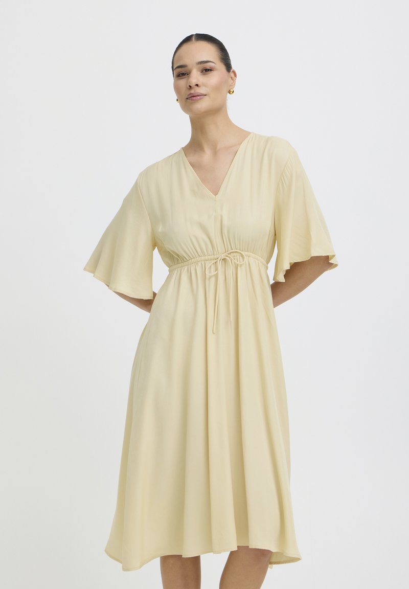 Femme portant une robe midi jaune clair avec des manches volantées et une taille à cordon, debout, les mains derrière le dos, sur fond blanc.