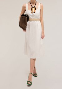 Witte jurk tot op de knie met een vierkante halslijn, een getailleerde taille en een beige riem. Groene sandalen met banden en een gevlochten bruine handtas maken de look compleet.