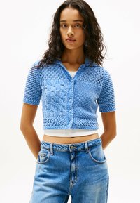 Gehaakte cardigan met korte mouwen in lichtblauw met een kabelgebreid patroon, knoopsluiting en textuurdetails; gedragen over een wit top.