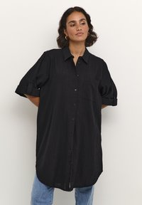 Chemise noire surdimensionnée avec des manches courtes retroussées, boutonnée à l'avant, poche poitrine unique et tissu texturé, portée sur un jean bleu clair.