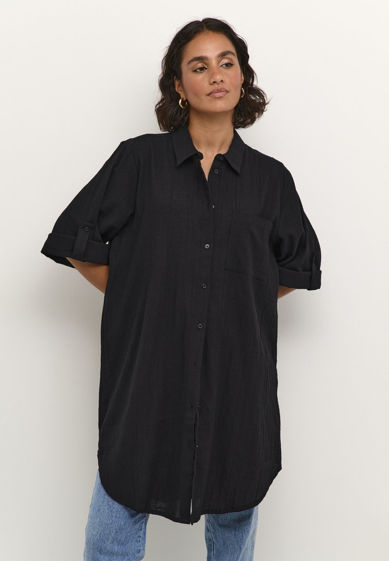 Chemise noire surdimensionnée avec des manches courtes retroussées, boutonnée à l'avant, poche poitrine unique et tissu texturé, portée sur un jean bleu clair.