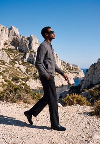 Homme portant des lunettes de soleil, un pull gris et un pantalon noir marchant sur un sentier rocheux avec des falaises et la mer bleue en arrière-plan sous un ciel dégagé.