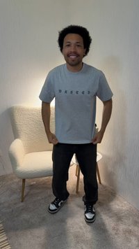 Άνδρας με σγουρά μαλλιά που χαμογελάει, φοράει γκρι μπλουζάκι με τη λέξη «WASTED», μαύρο παντελόνι και αθλητικά παπούτσια, στέκεται σε ένα χαλιεμένο δωμάτιο με πολυθρόνα και μικρό τραπέζι.