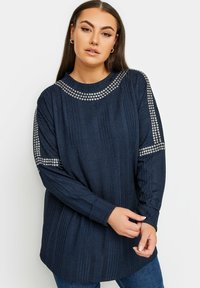 Marinblå långärmad topp med ribbad struktur, dekorerad med silverfärgade nitar längs axlarna och halsringningen. Lätt lös passform med rund halsringning.