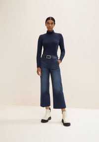 Marinefarbener Rollkragenpullover, hochgeschnittene, dunkelblaue, weite Jeans und weiße Knöchelstiefel mit schwarzer Sohle; ausgestattet mit einem Gürtel mit silberner Schnalle.