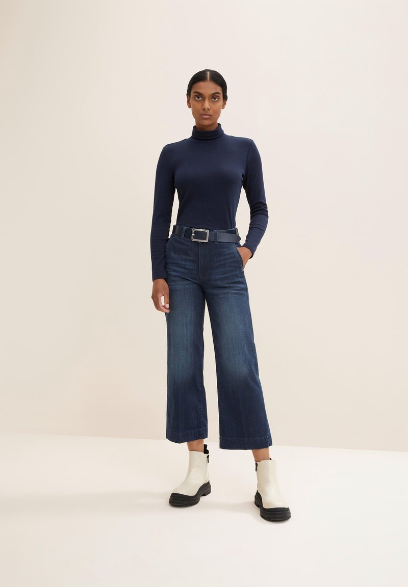 Marinefarbener Rollkragenpullover, hochgeschnittene, dunkelblaue, weite Jeans und weiße Knöchelstiefel mit schwarzer Sohle; ausgestattet mit einem Gürtel mit silberner Schnalle.