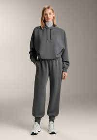 Szürke kapucnis pulóver magas nyakkal, húzózsinórral és oldalzsebekkel, hozzáillő szürke melegítőnadrággal, bordázott mandzsettákkal és laza szabással.
