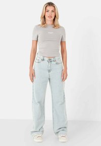 Grijze fitted t-shirt met korte mouwen en wit logo, gecombineerd met lichtblauwe wijde jeans en off-white sneakers. Eenvoudige, casual outfit.